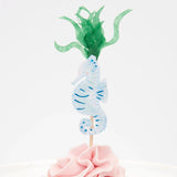 A light blue seahorse cupcake topper with green seaweed fronds on a pink frosted cupcake.
