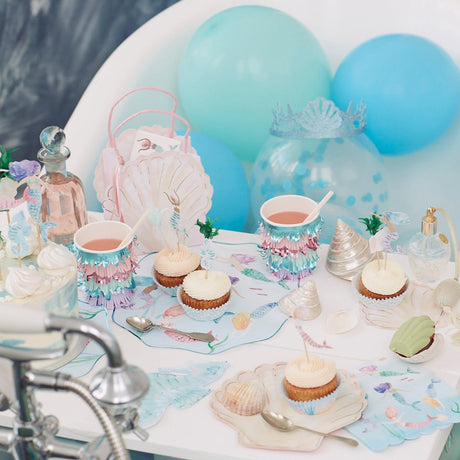 Mermaid-themed party table with cupcakes, decorated cups, balloons, and clam shell tableware.
