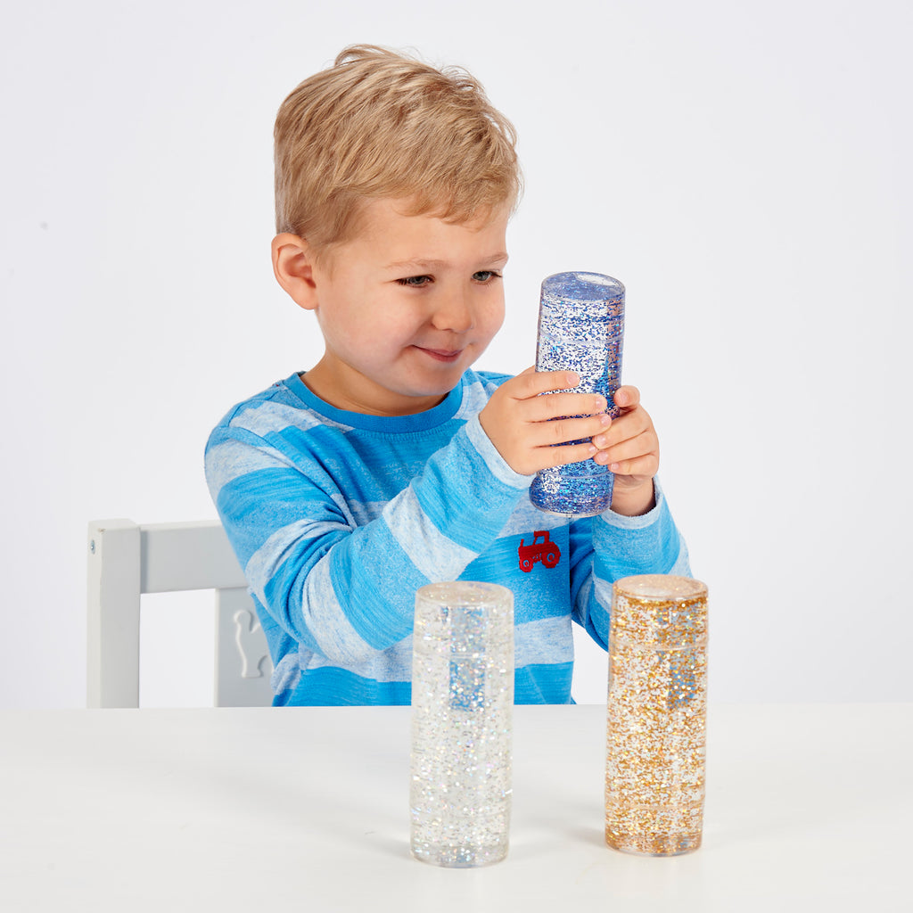 A child in a blue striped shirt is smiling and holding a clear cylinder filled with blue glitter. Two similar glitter cylinders are on a white table.