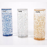 Three tall, clear cylinders filled with blue, silver, and gold glitter, set against a white background. Each has a black logo near the top.