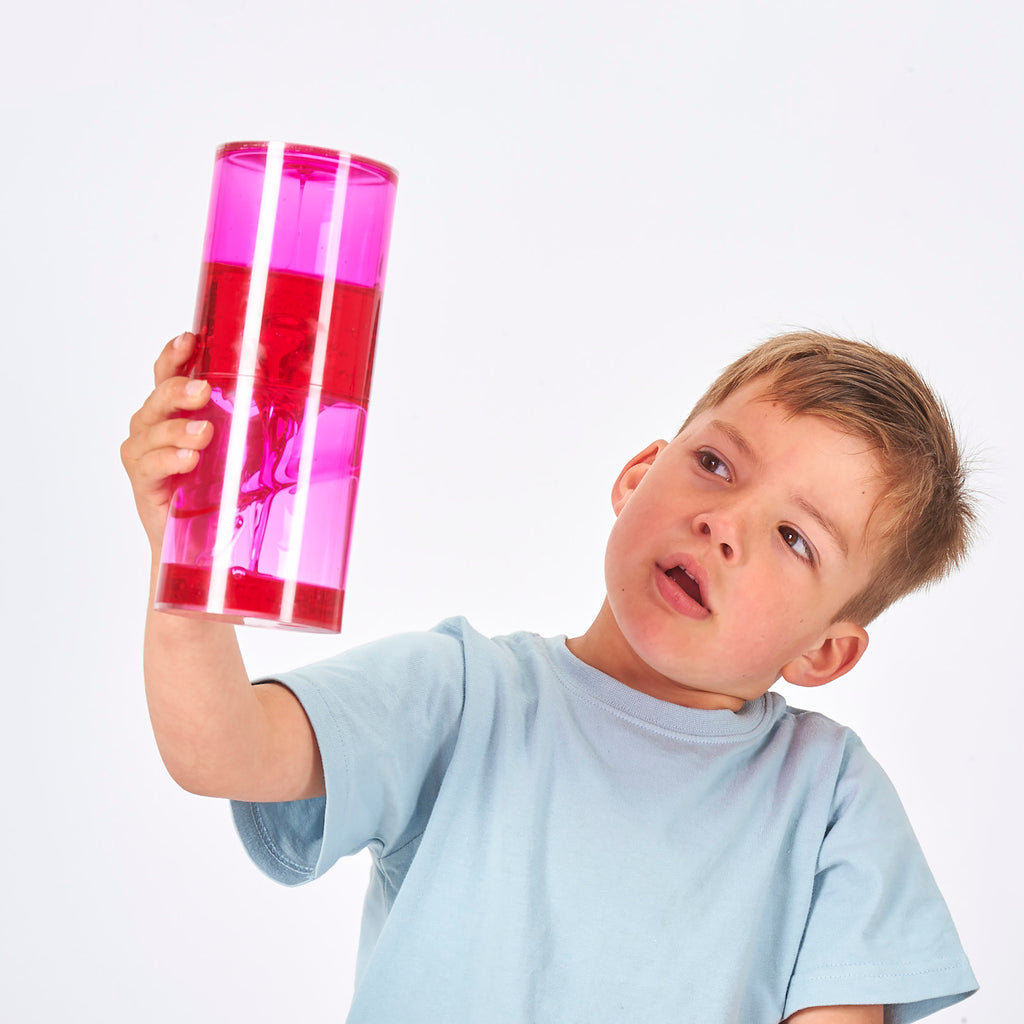 A young boy in a light blue shirt curiously examines a pink and red lava lamp held up in his right hand. The white background highlights his focused expression.