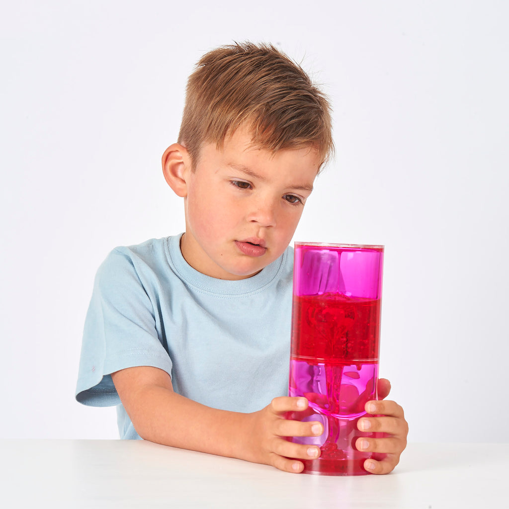 A young boy in a light blue shirt curiously examines a pink and red lava lamp held up in his right hand. The white background highlights his focused expression.