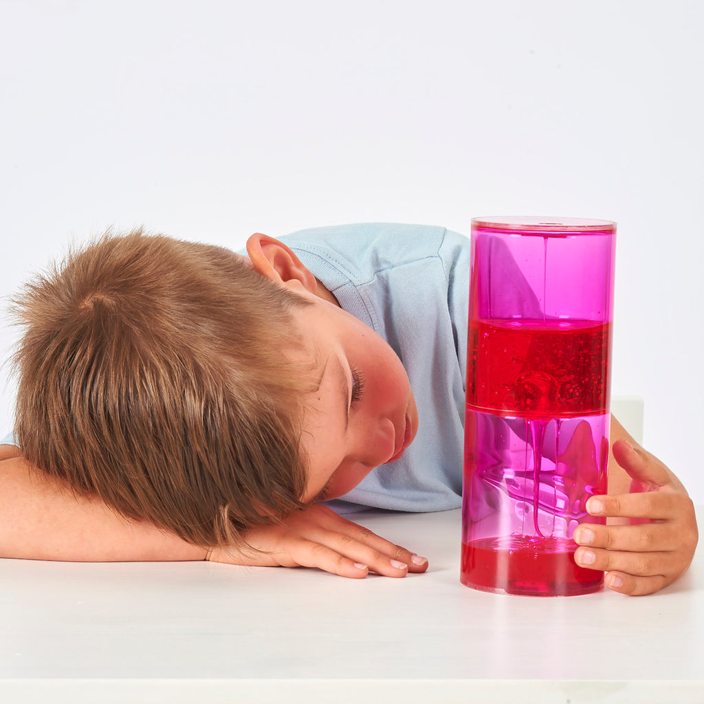 A young boy in a light blue shirt curiously examines a pink and red lava lamp held up in his right hand. The white background highlights his focused expression.