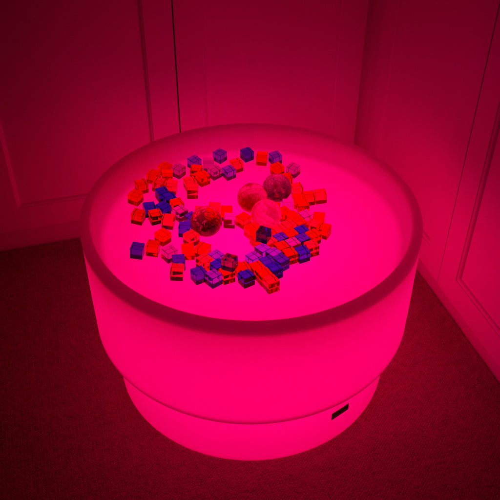 A round table bathed in vibrant pink light, scattered with colorful toy blocks and several textured balls. The scene is playful and surreal.