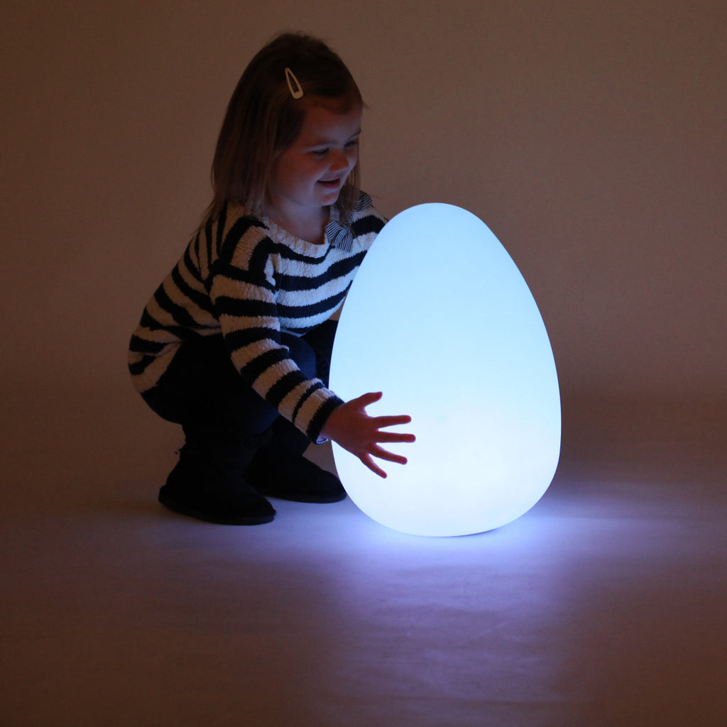 A child, wearing a black and white striped sweater, happily crouches beside a glowing egg-shaped lamp, casting a soft blue light in a dim room.