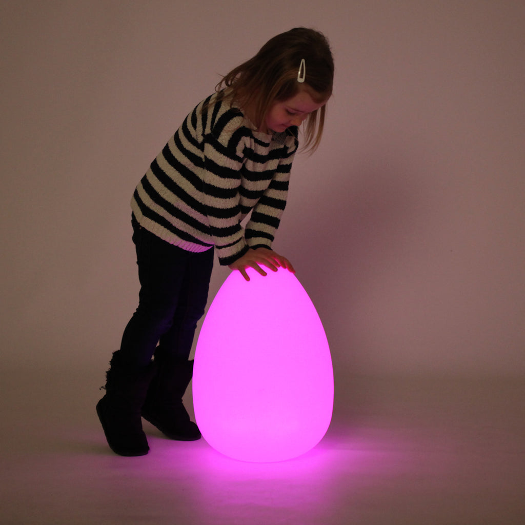 A child, wearing a black and white striped sweater, happily crouches beside a glowing egg-shaped lamp, casting a purple light in a dim room.
