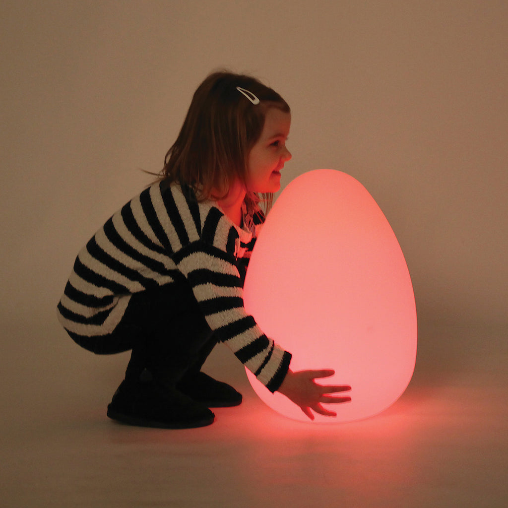 A child, wearing a black and white striped sweater, happily crouches beside a glowing egg-shaped lamp, casting a soft red light in a dim room.