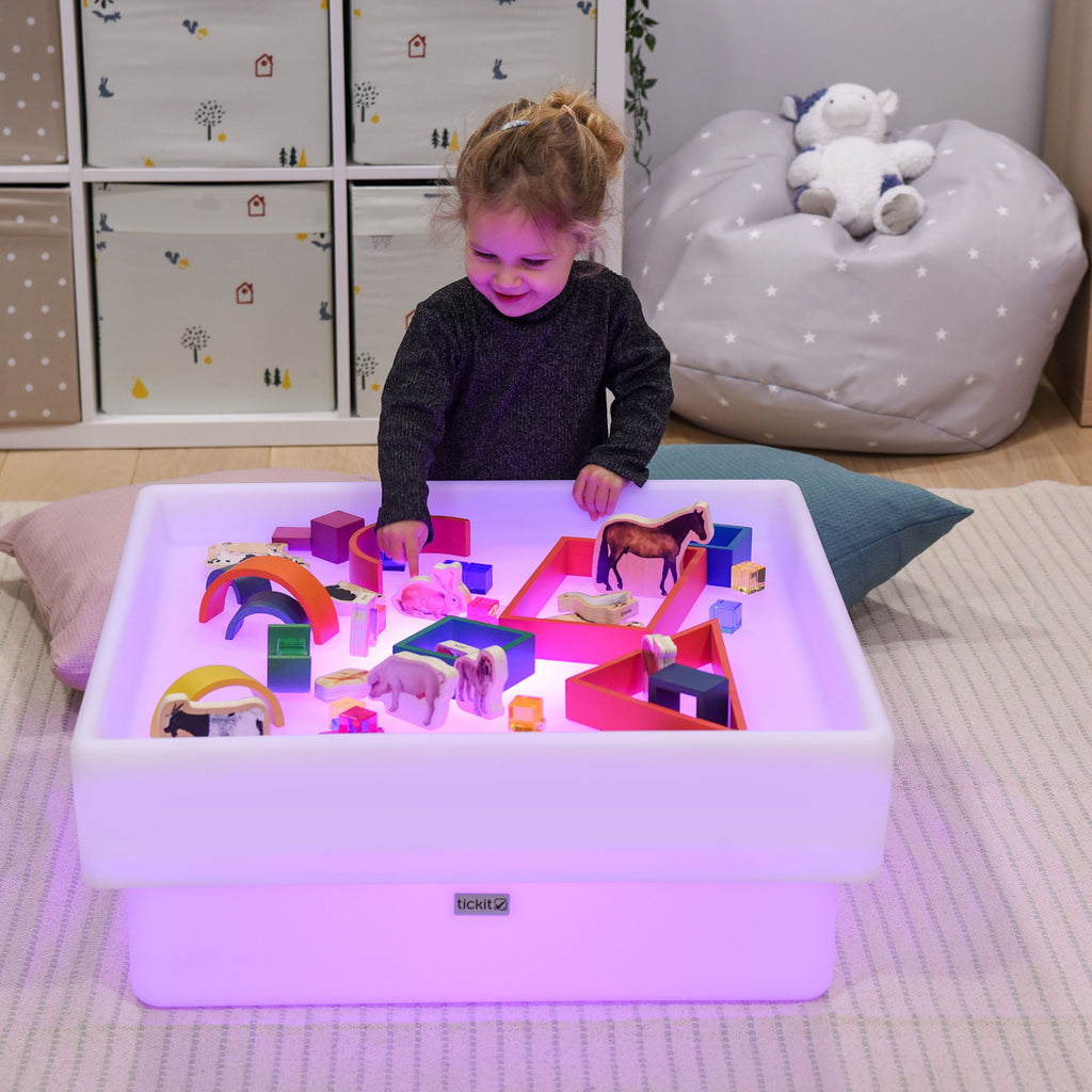 A child smiles while playing with colorful toy animals and blocks on a glowing light table. The room is cozy, with storage boxes and a beanbag.