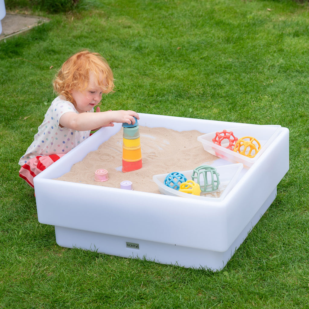 A child plays in a white plastic sandbox on a grassy lawn, stacking colorful cups. Bright toy balls are placed in a container nearby, conveying a playful mood.
