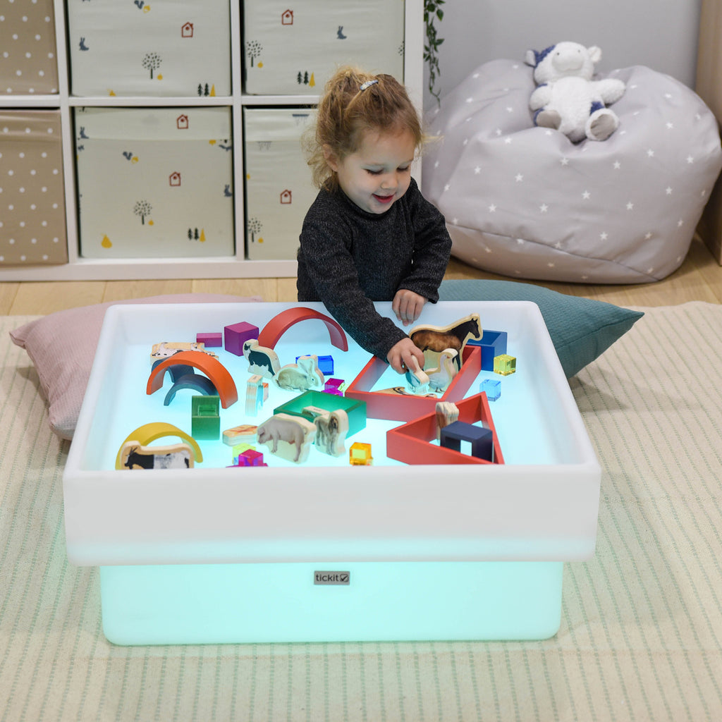 A child smiles while playing with colorful toy animals and blocks on a glowing light table. The room is cozy, with storage boxes and a beanbag.