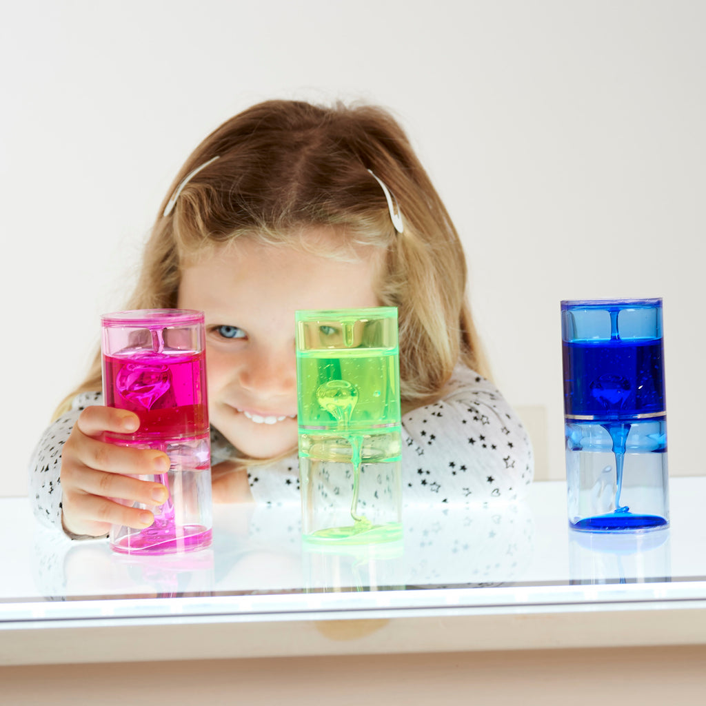 A smiling child with long hair holds a pink liquid timer, surrounded by green and blue timers on a reflective surface. The scene feels playful and colorful.