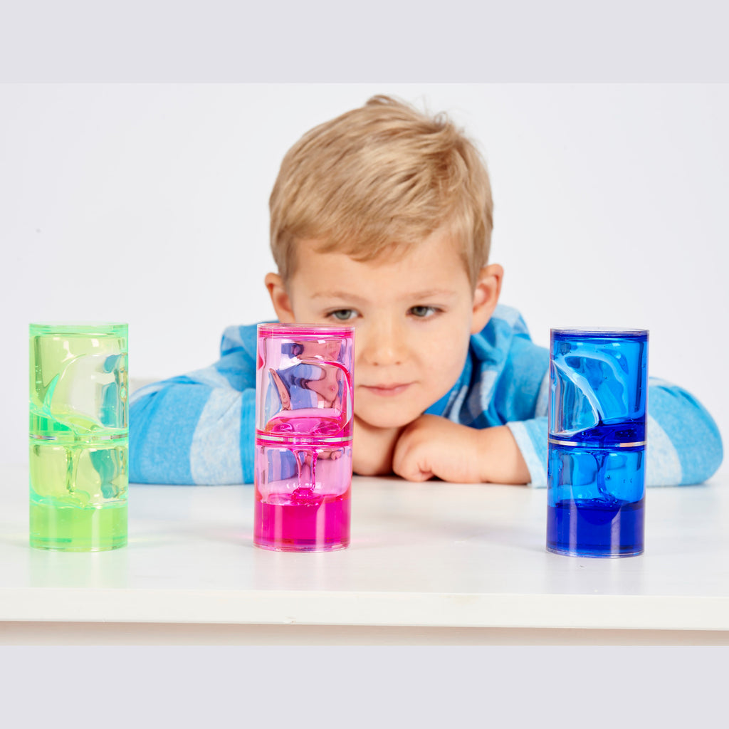 A young child in a blue-striped shirt looks intently at three colorful liquid timers on a table. The timers are green, pink, and blue, conveying curiosity.