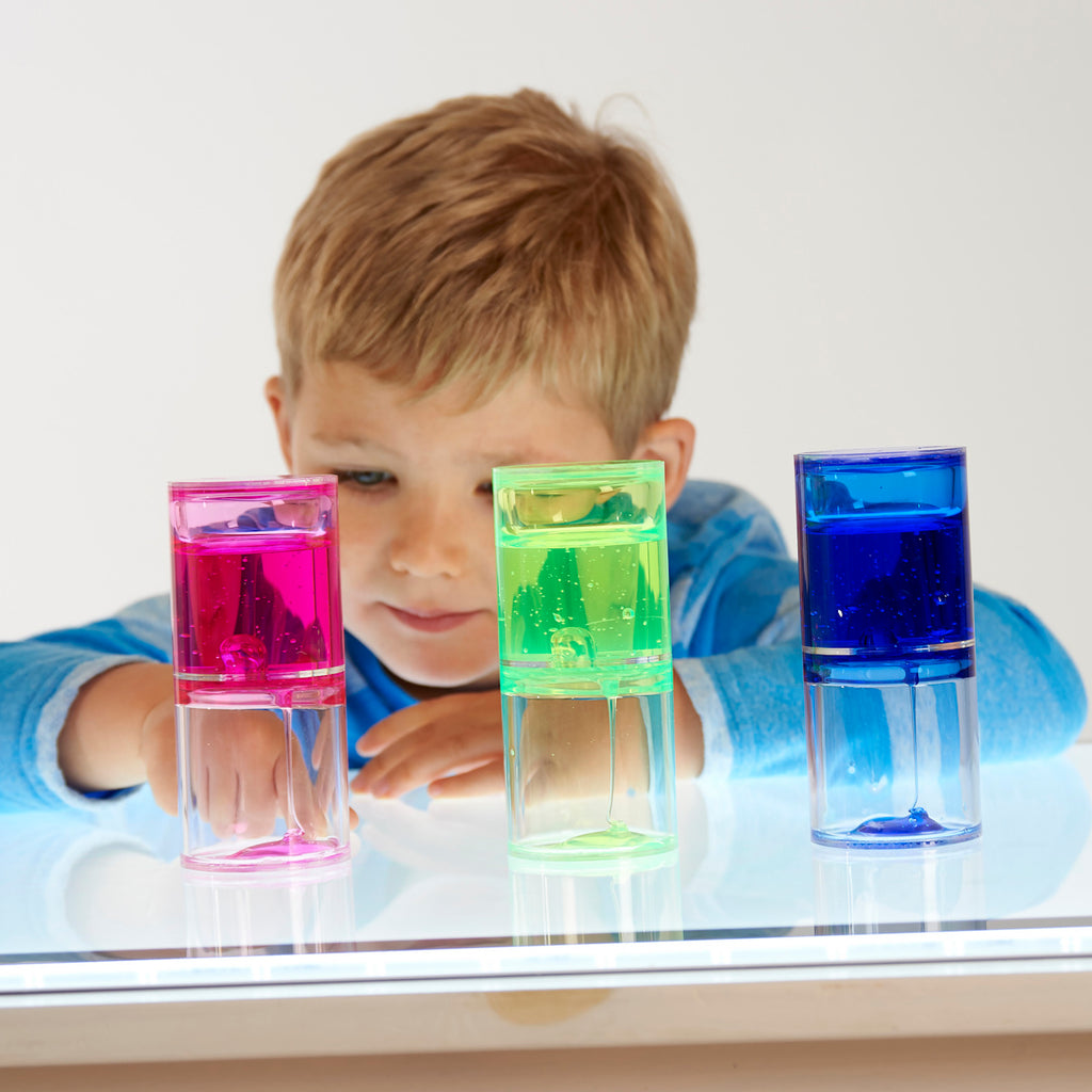 A young child in a blue-striped shirt looks intently at three colorful liquid timers on a table. The timers are green, pink, and blue, conveying curiosity.