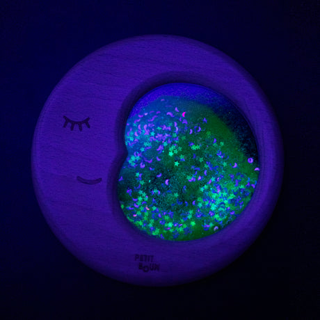 A Petit Boum Flow Moon sensory toy glowing under UV light, showcasing sparkling stars and crescent moons in a dark setting.