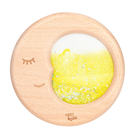 A close-up of the Petit Boum Flow Moon sensory toy, a wooden crescent filled with shimmering yellow glitter and glow-in-the-dark shapes.