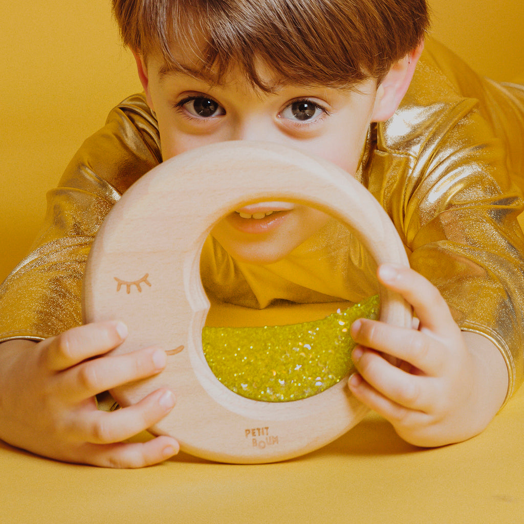 A child dressed in gold holding the Petit Boum Flow Moon sensory toy, engaging with its glittery, crescent-shaped sensory window.