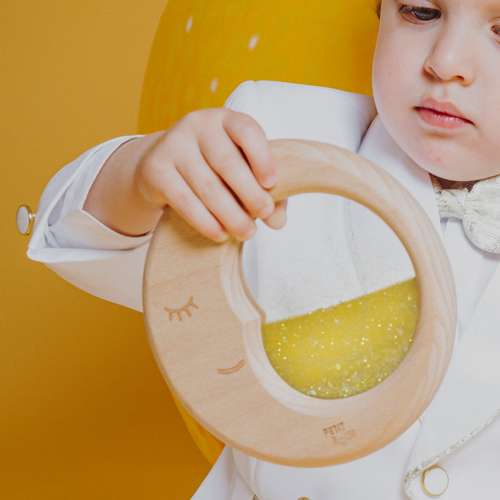 A small hand lifting the Petit Boum Flow Moon sensory toy against a yellow background, showing off the dreamy crescent moon design.