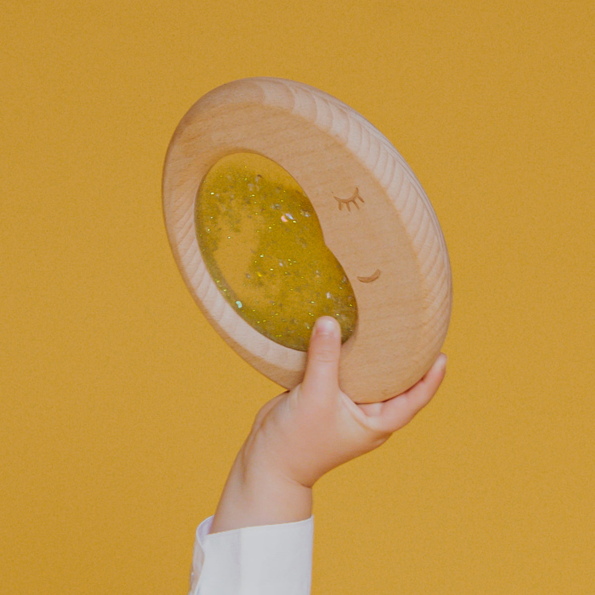 A small hand lifting the Petit Boum Flow Moon sensory toy against a yellow background, showing off the dreamy crescent moon design.