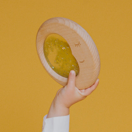 A small hand lifting the Petit Boum Flow Moon sensory toy against a yellow background, showing off the dreamy crescent moon design.