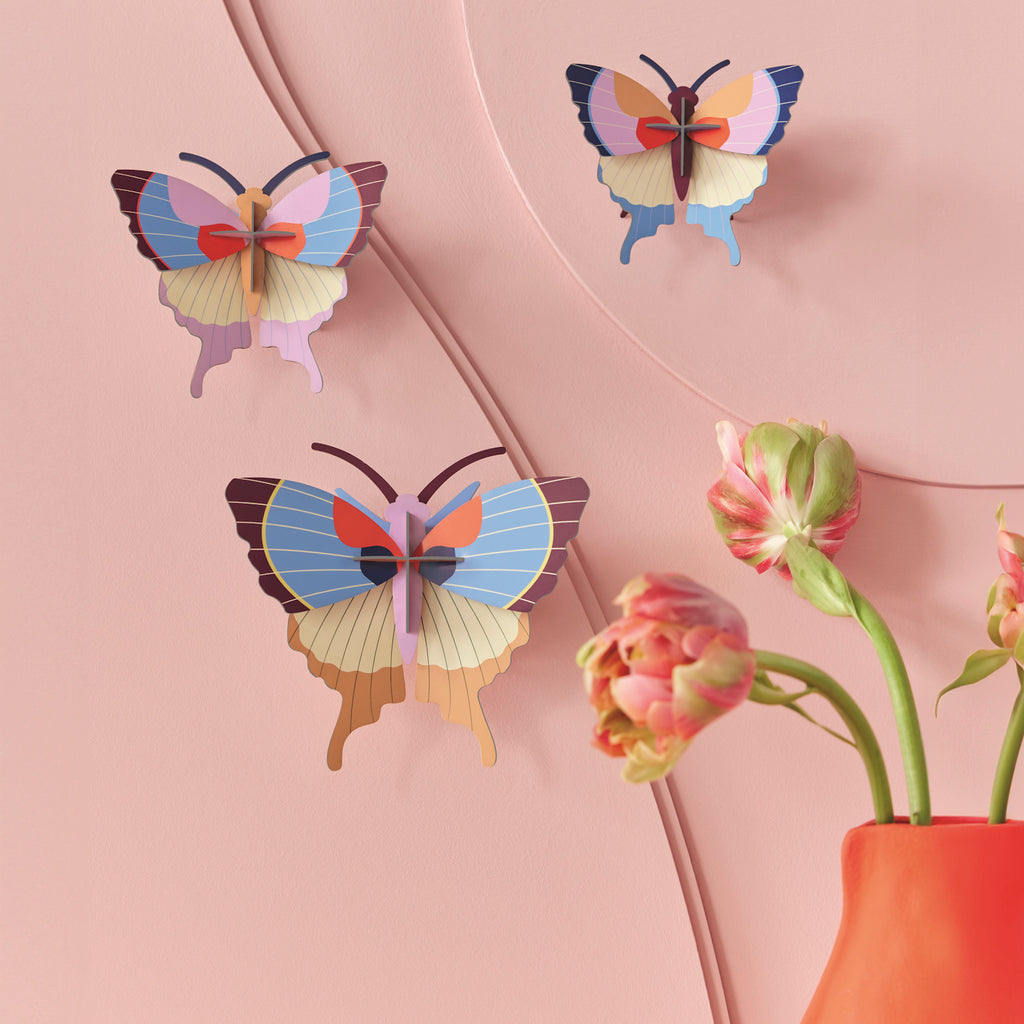 Three colorful paper butterflies are mounted on a pastel pink wall near a red vase with pink tulips. The scene is bright and whimsical.