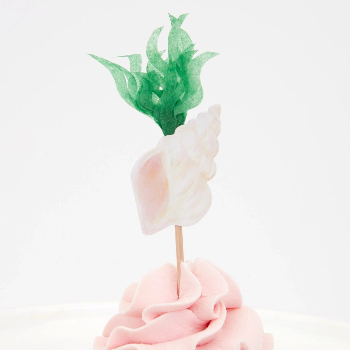 A cupcake with pink frosting, topped with a decorative seashell and green seaweed paper topper.