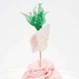 A cupcake with pink frosting, topped with a decorative seashell and green seaweed paper topper.