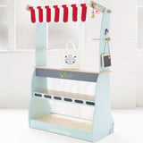 Wooden toy market stall with striped awning, shelves, shopping bag, and chalkboard in a room.
