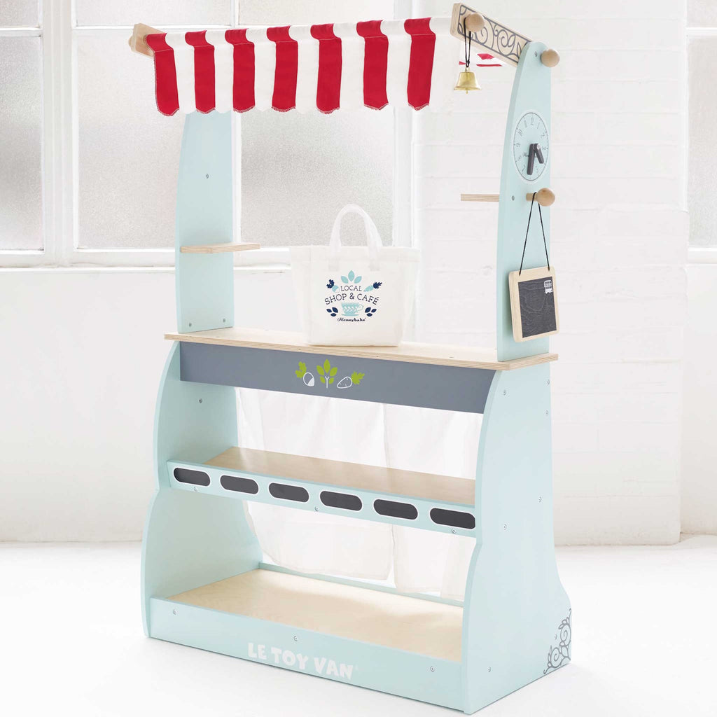 Toy market stall with red and white striped canopy and open sign. Features shelves labeled with food categories, a bell, and a hanging bag. Bright and playful.