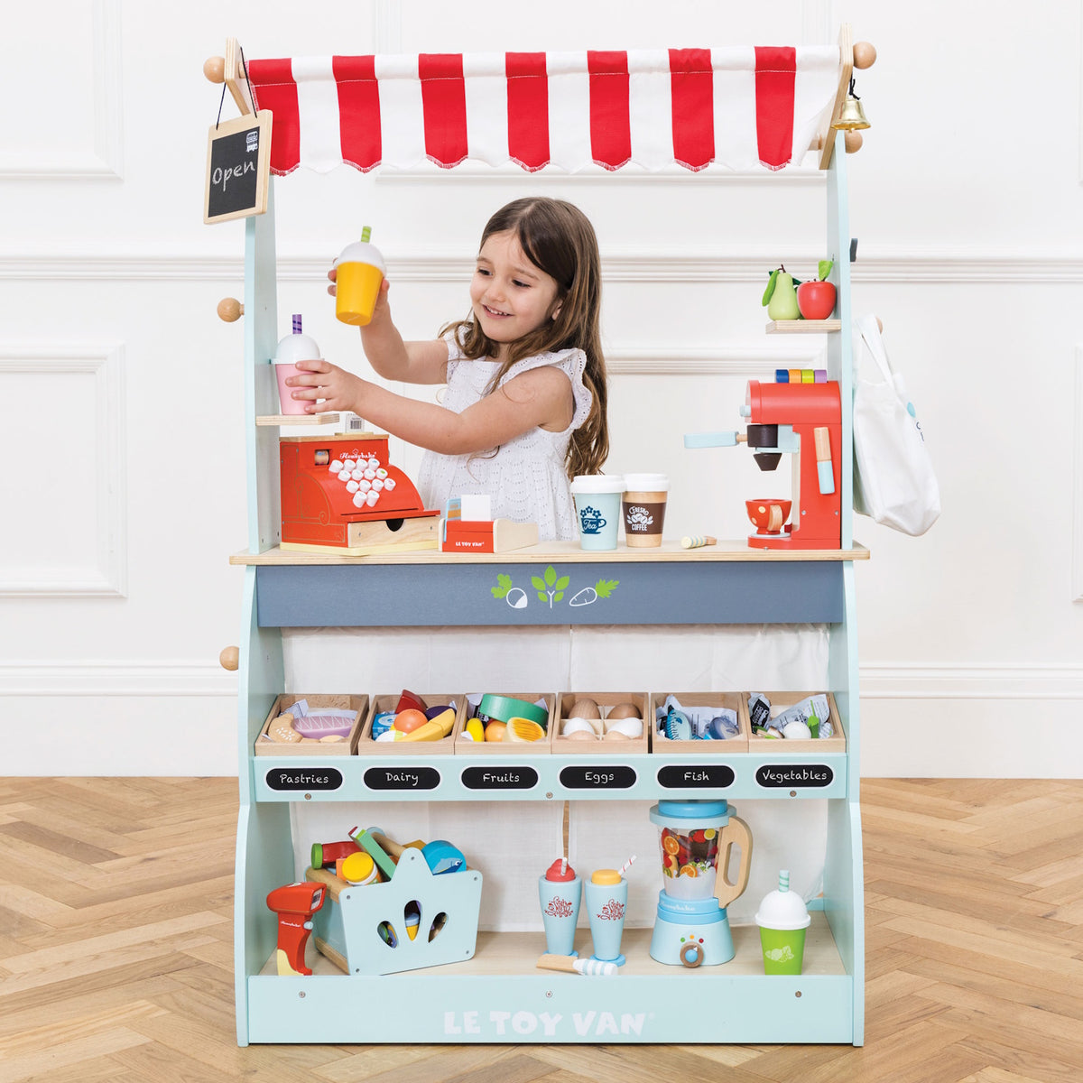 Young girl playing behind a fully stocked wooden market stand and café toy set.