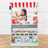 Young girl playing behind a fully stocked wooden market stand and café toy set.