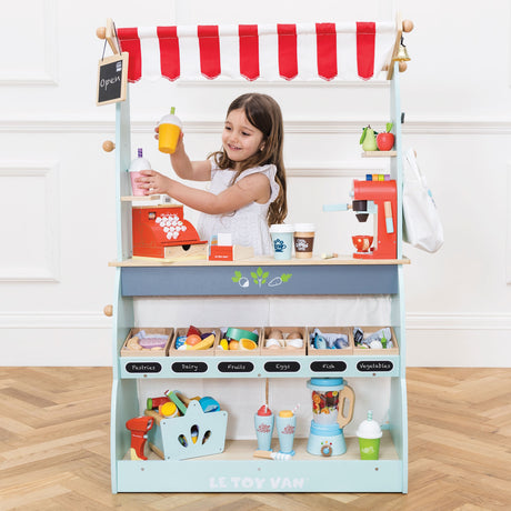 Young girl playing behind a fully stocked wooden market stand and café toy set.