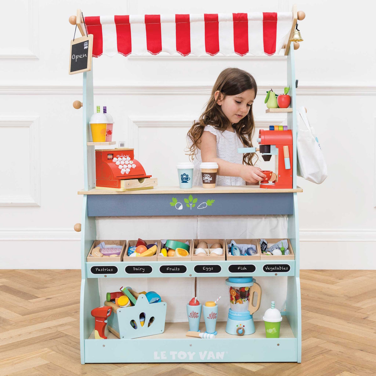 Young girl playing behind a fully stocked Le Toy Van wooden market stall and café set on a herringbone floor.