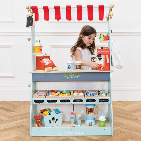 Young girl playing behind a fully stocked Le Toy Van wooden market stall and café set on a herringbone floor.