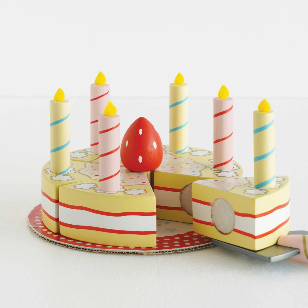 Toy cake with yellow layers, white frosting, and red stripes, topped with colorful candles and a red strawberry. A slice is on a spatula. Playful tone.