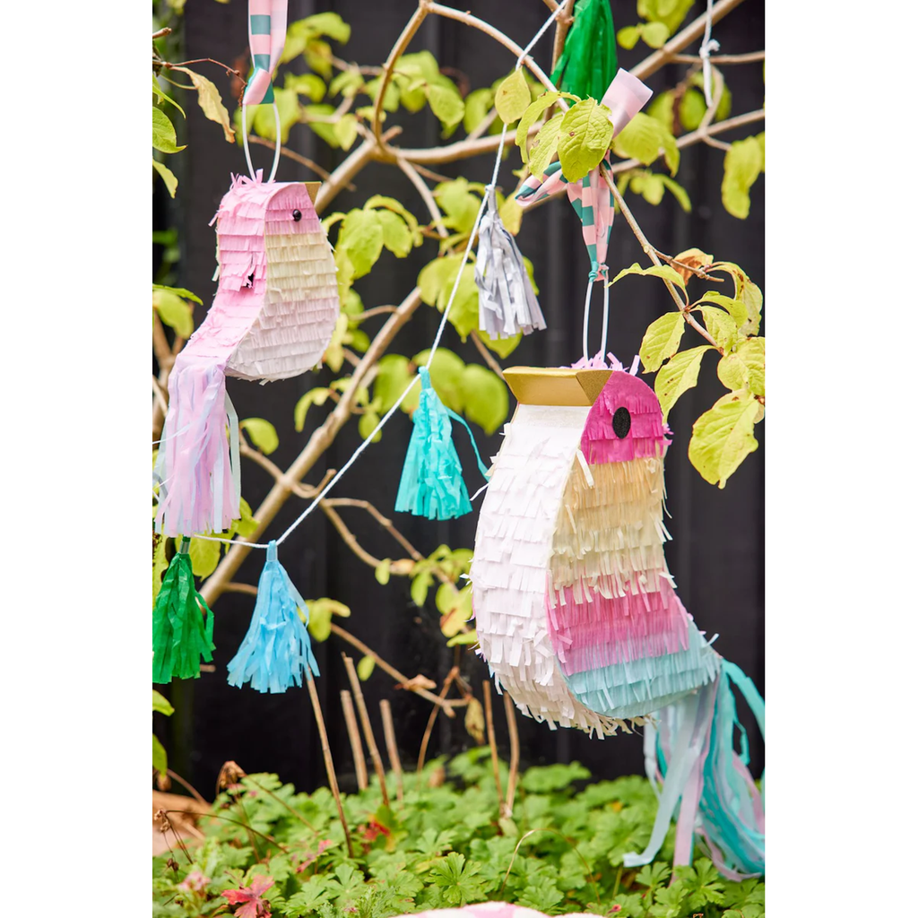 Colorful bird-shaped piñatas hang among green leaves, adorned with pastel tassels in pink, blue, and green, creating a festive, whimsical atmosphere.