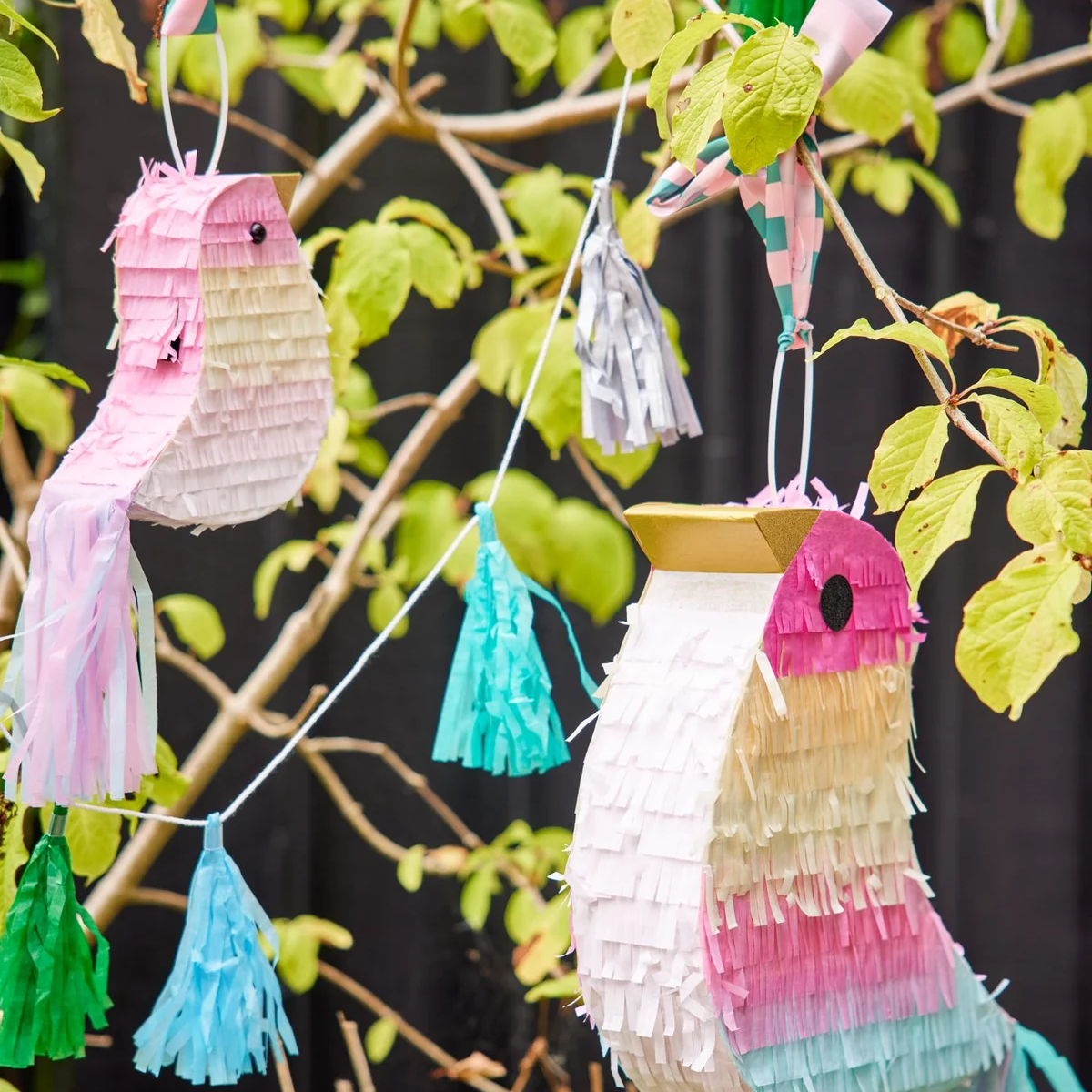 Colorful bird-shaped piñatas hang among green leaves, adorned with pastel tassels in pink, blue, and green, creating a festive, whimsical atmosphere.
