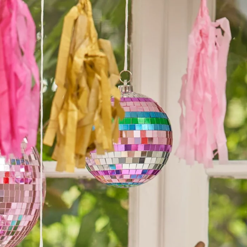 Colorful disco balls, tassels, and paper fans hang by a window with a bird vase and flowers. Vibrant and festive atmosphere.
