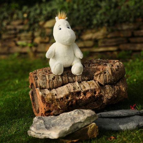A plush white hippo toy with a tuft of yellow hair sits on aged logs in a garden, surrounded by green grass and a stone wall background, conveying a whimsical outdoor scene.