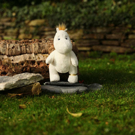 A white plush toy with an orange tuft of hair sits on a stone slab in a garden. It is surrounded by mossy stones and greenery, exuding a peaceful vibe.