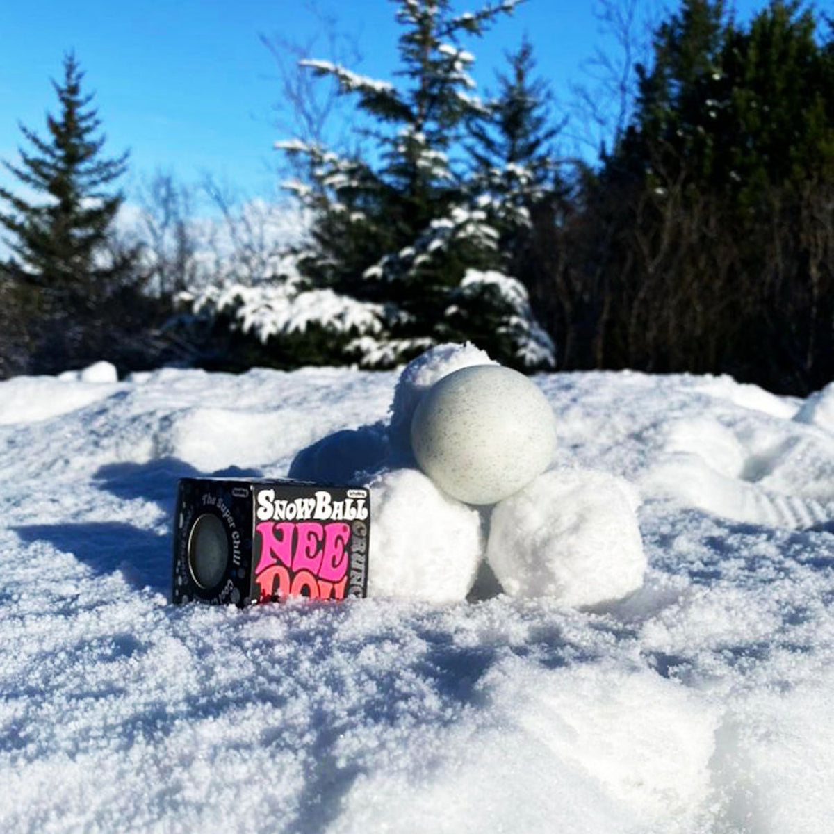 Schylling Snow Ball Crunch NeeDoh stress ball sitting on real snow next to its packaging with pine trees in the background.