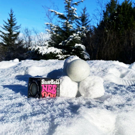 Schylling Snow Ball Crunch NeeDoh stress ball sitting on real snow next to its packaging with pine trees in the background.