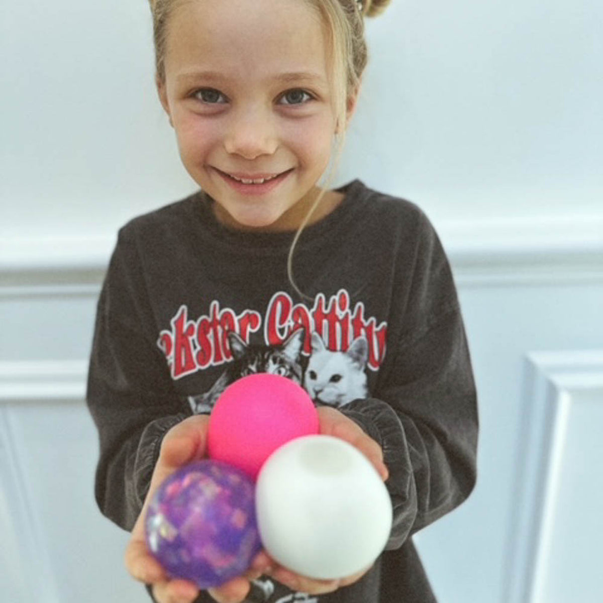 Smiling young girl holding pink, purple, and white snowball crunch stress balls while wearing a cat graphic sweatshirt.