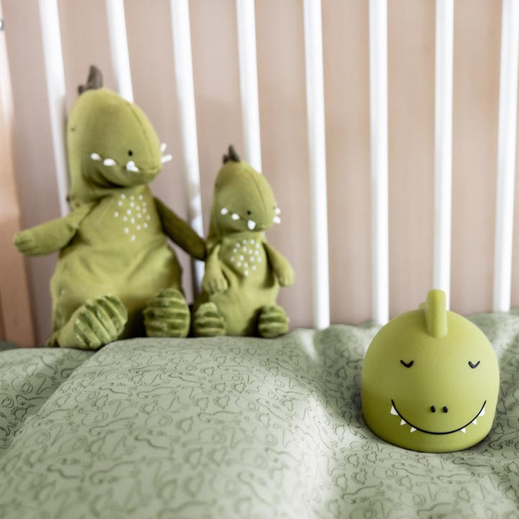 Two green dinosaur plushies and a dinosaur night light sit on a crib with a soft, patterned sheet. The scene conveys a cozy, playful atmosphere.
