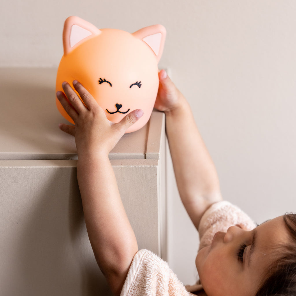 A child reaches for a glowing cat-shaped night light on a table. The light is orange with a smiling face, conveying a warm and playful atmosphere.