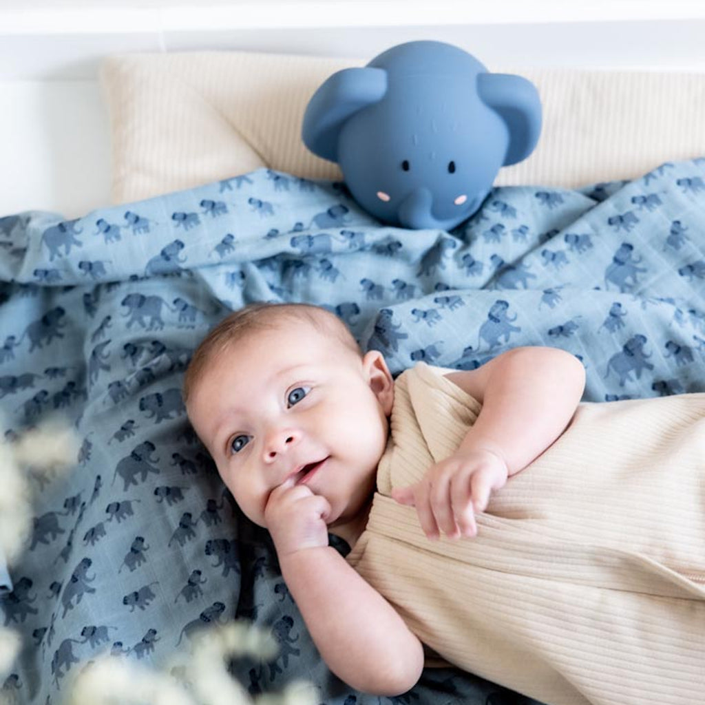 A smiling baby lies on a blue blanket with elephant patterns, wearing a beige outfit. A blue elephant toy is in the background, creating a cozy, playful mood.