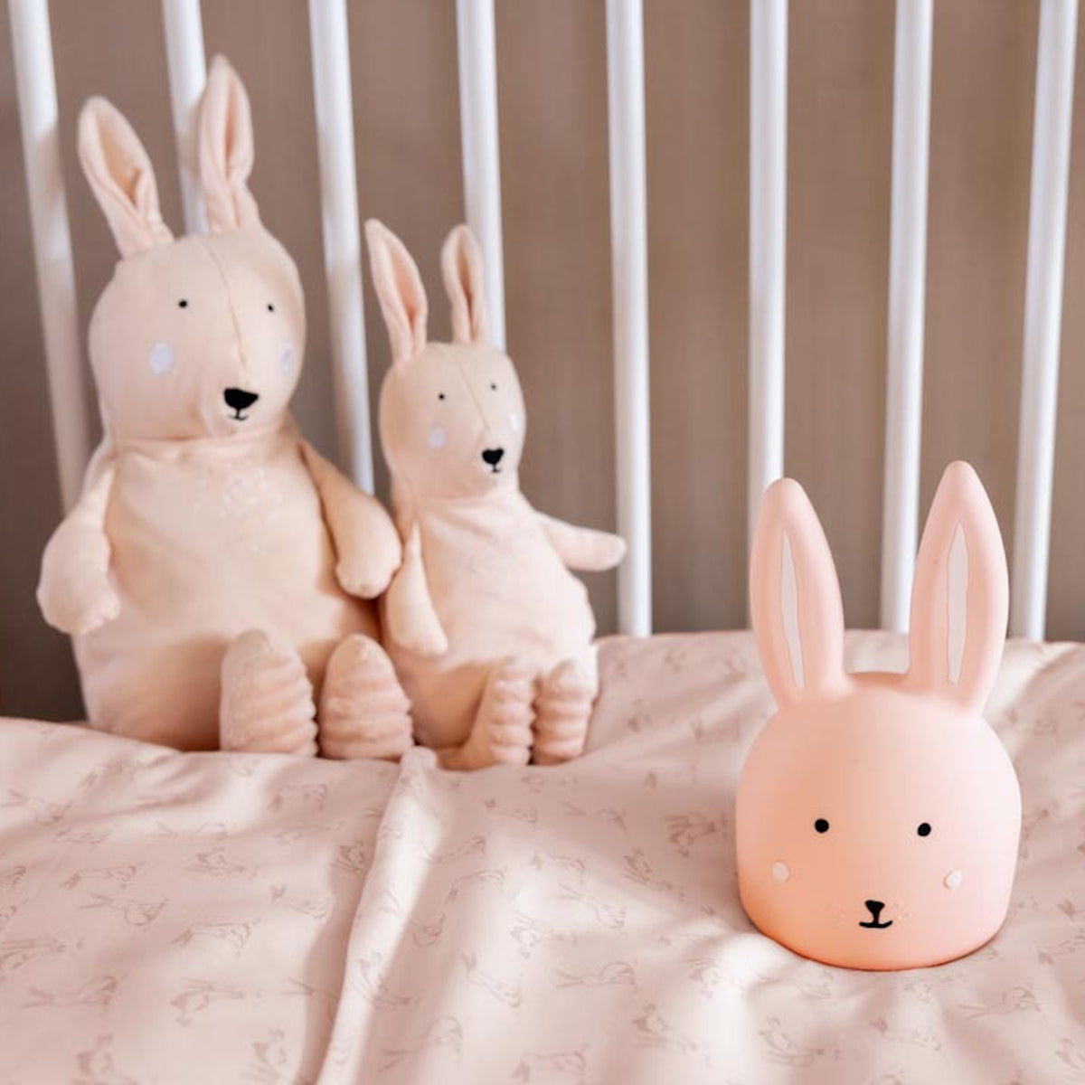 Stuffed bunny toys and a soft bunny lamp sit on a crib mattress with pale bedding. The scene is cozy and serene, emitting a warm, gentle vibe.