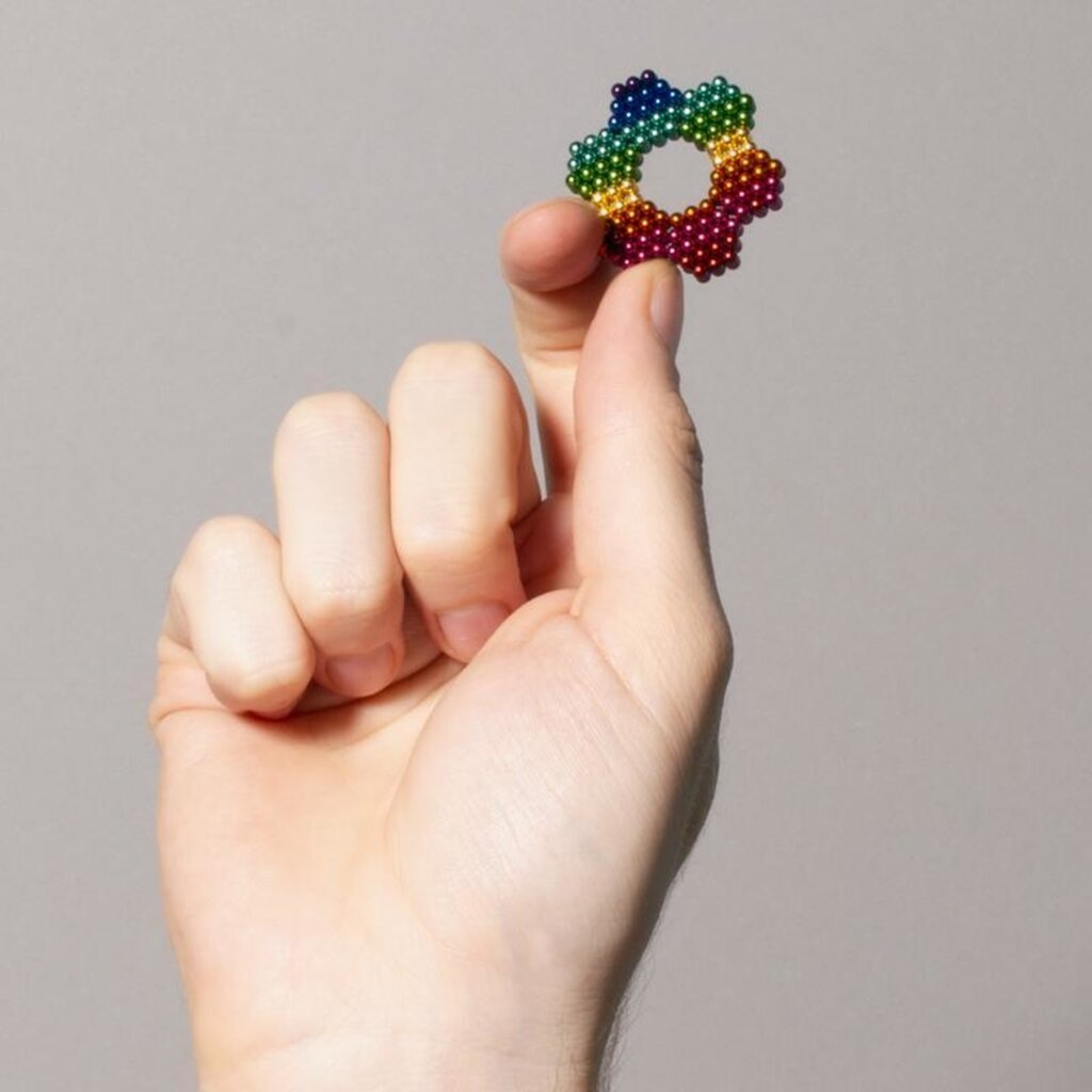 A hand holding a small circle made of colorful magnetic balls, arranged in a rainbow pattern. The background is a plain, neutral gray.