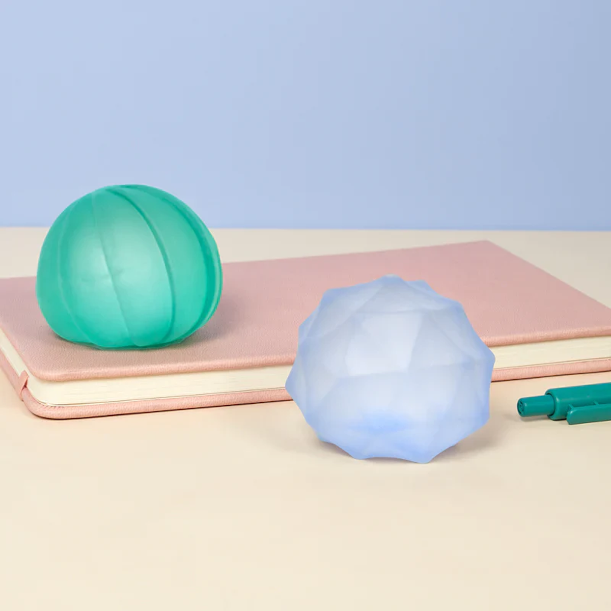 Two stress balls, one green and one faceted blue, rest on a pink notebook. A green pen is partially visible. The scene has a calm and minimalist tone.