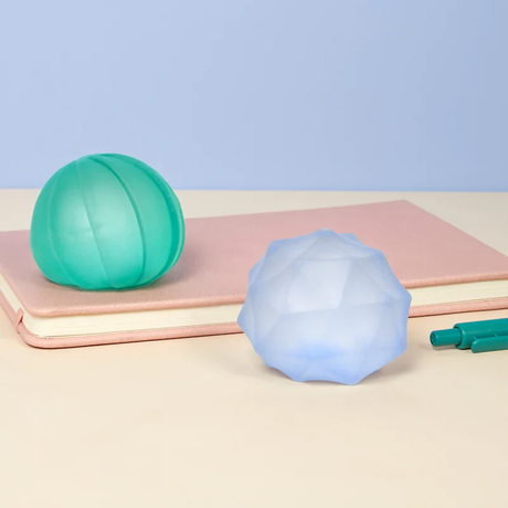 Two stress balls, one green and one faceted blue, rest on a pink notebook. A green pen is partially visible. The scene has a calm and minimalist tone.