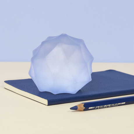 A geometric, translucent blue orb rests on a closed blue notebook, next to a blue colored pencil. The scene conveys calm and creativity.